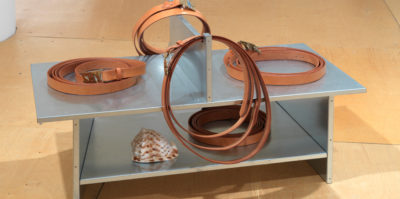 A photograph of a metal sculpture. The metal sculpture is situated on the floor, and takes the form of a small, two-layer table. On the lower shelf, there is a white and brown sea shell and a brown belt. On the top of the piece, there are four more brown belts on each side of the metal sculpture.