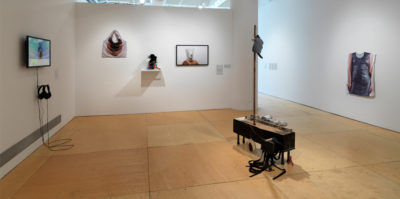 An installation photograph from inside square, gallery space. On the left wall, a television monitor is mounted against the wall with headphones. The film still on the monitor is of a person standing in front of a blue and green background. On the center wall, three objects are mounted on the wall. The object on the left is a white, brown and black layered textile that is in the shape of a square with a handle. The center object is placed on a white shelf. The object on the right is of a portrait photograph of a person wearing a white mask. On the right wall there is an image of a person wearing a black dress. In the center of the entire space there is a rectangular sculpture standing with four pegs on the floor. There is a tall wooden line extending from the base. On the right side of the sculpture, six aluminum cans are situated horizontally.