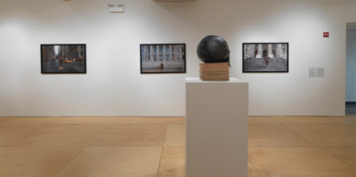 An installation photograph of three large, framed industrial landscape photographs mounted on a wall. In front of the wall, there is a white, tall square podium with eight, medium sized, light brown books stacked vertically. Sitting on the stack of books is a black, circular object about the size of basketball.The circular object has black hair attached to its back side.
