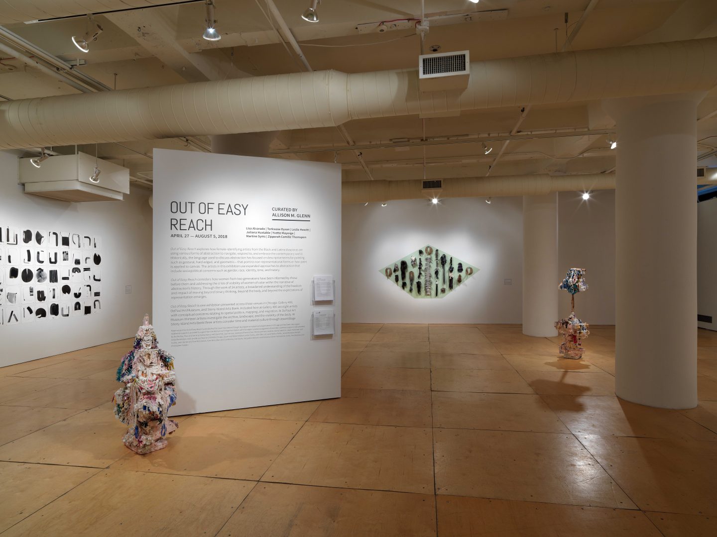 An Installation photograph of the gallery. A separate, square column stands on its own in the center of the photograph. On the column is adhered text of the exhibition information. The title, in large, bold texts, writes, “Out of Easy Reach”. On another wall to its left is a large black and white print of multiple different shapes. On another wall to its right is a pained, large, green rhombus. Adhered to the rhombus are various types of hair yarn, and materials.