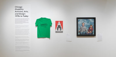 An installation photograph of the gallery wall. On the left of the wall, the introductory text to the gallery is pasted. Four paragraphs of small text are vertically situated. Larger, bolded text at the top of the paragraphs is the exhibition title that writes, “Chicago Disability, Activism, and Design: 1970’s to today. On the right of the text is a green t shirt, a poster, and a painting of two people in a black frame.