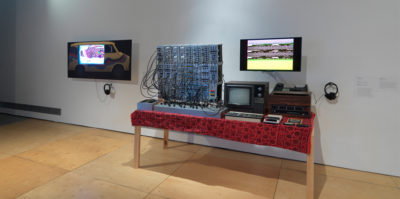 An installation photograph of the main gallery’s right side wall. Two television monitors are mounted against the wall. Below the television monitor on the right side of the photograph, is a six foot long wooden table. On the table sits a red and maroon printed table cloth. Sitting on the table is a small, 80’s television with wood detail and a large synthesizer, about twice the size, with multiple cords.