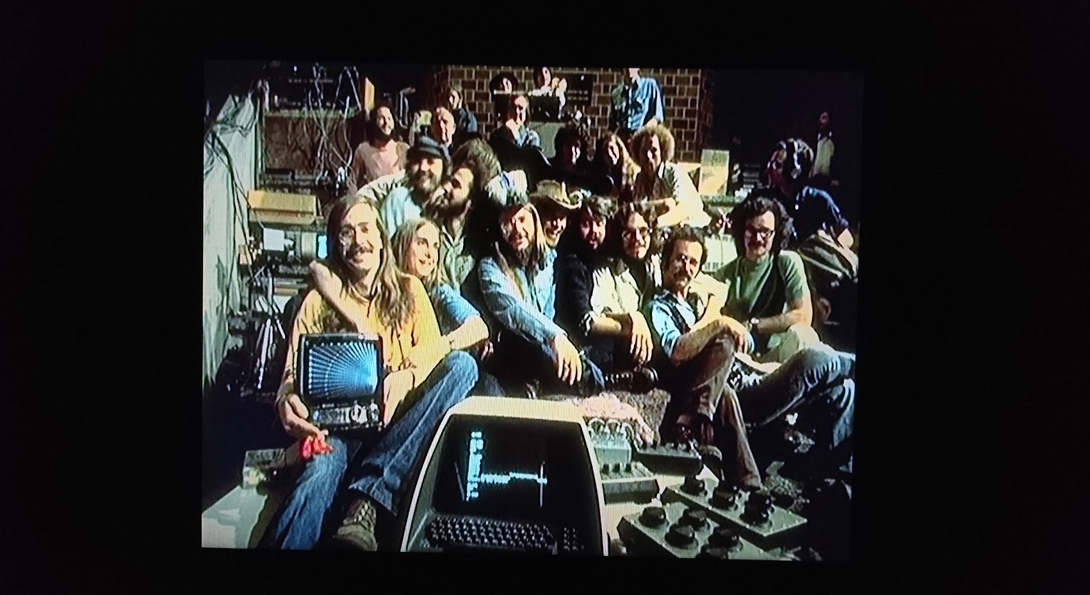 A photograph of fifteen people in a group, sitting closely. They looking at the camera, smiling, and holding 1970s computer softwear.