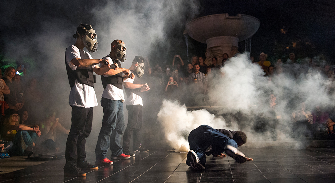 A photograph of a performance, taken at night-time. A group of spectators are circled around them as they perform outside, under a light. Three performers are standing in a line with gas masks on and bullet-proof vests. There is a smoke effect behind them. They are wearing jean pants and white T-shirts. There is a person on the ground in front of them.