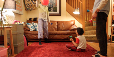 A photograph of a video still from Sensitive Equipment. The video still is in a living room setting. A young person is sitting on a red carpet on the floor. They are looking upwards at another young person, standing, holding a pink balloon. They are both wearing pajamas and engaging in front of a brown leather couch. Behind the couch, there are wooden stairs leading up to the top center of the photograph. On the right side of the photograph, the lower body of an older person is standing with toys in their hand.