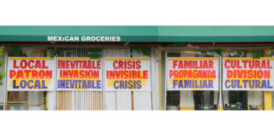 A long, landscape photograph of a mexican grocery store. Five window signs have bolded text in red, orange, blue and yellow, printed on white poster paper. The sign in the center window reads, “CRISIS. INVISIBLE. CRISIS”. Above the windows is a green rooftop that reads, “Mexican Groceries”.