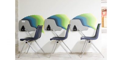 A photograph of three plastic and metal, blue school chairs are positioned against a white wall. The top of the desk, connected to each chair, is flipped upward against the wall. A design takes the shape of each of the chair’s desks and is painted in a gradient from blue, green, and then yellow.