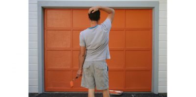 Photograph of a middle-aged person standing in front of an orange garage door. The person, wearing gray clothing, is holding an orange roller paint brush. Beside them, a tin of orange paint sits on the floor.