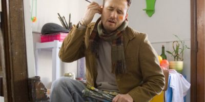 A photograph of a middle-aged person is sitting in a chair holding paint brushes. They are wearing a brown jacket, jeans, and a brown scarf. Behind them, there is furniture holding plans, paint brushes, and a white towel.. The person is holding a paintbrush, sticking it in their left ear, with orange paint on their right cheek. Closing their eyes, their face holds a displeased reaction.