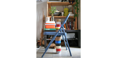 A digital, portrait-style photograph of a light blue, small, metal chair. The photograph is taken closely, so the chair fills up most of the frame. Books, an orange cooler, and a white coffee cup are stacked on top of the base of the chair. 13 rolls of electric tape, in multiple colors, are stacked below the chair, and continue to stack upwards from the base of the chair. Behind the chair, a wooden bookshelf holds multiple books, cups, and green plants.