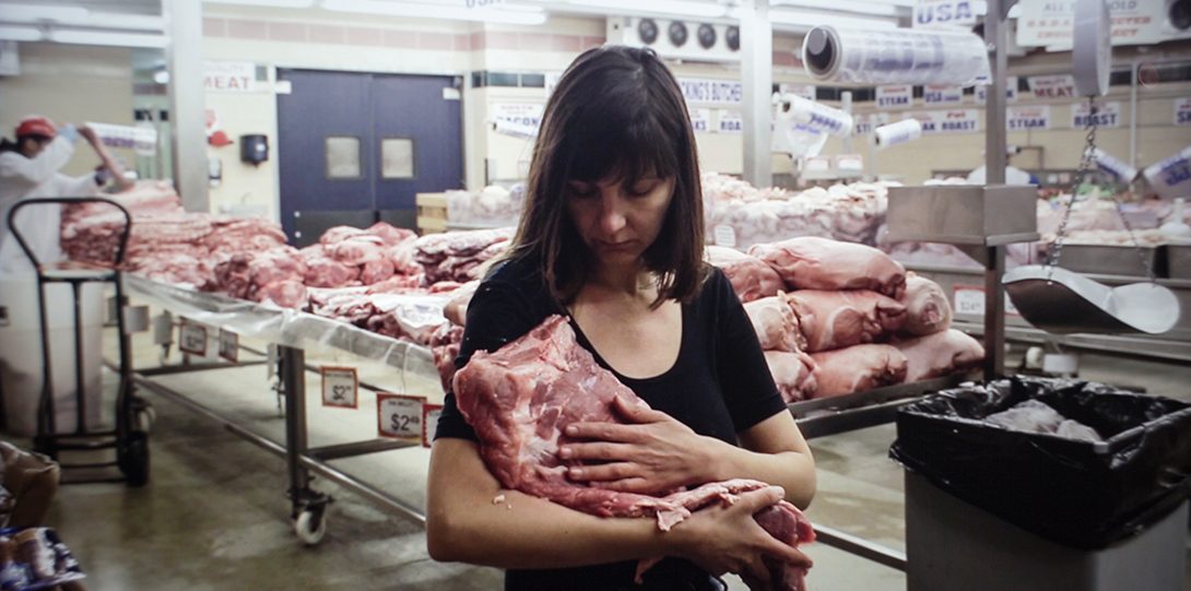 A photograph of a middle aged person holding a large, square piece of raw meat. They are holding it in their left arm, and their right arm is placed on the front side. The person is standing inside a meat producing facility. Behind them is a large table of stacked, multiple square pieces of raw meat. Another person, in a white sanitary suit is working on the table behind them.