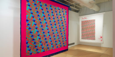 An Installation photograph of two large, ceiling to floor length, square textiles. The textile on the left has a thin, dark blue border with fringe draped from the bottom. Inside this border is another, much thicker, dark pink border. Inside is a zig-zag design of small squares in blue, red, teal, and orange colors. The textile on the right has a thick, white border. The design inside the border is a laced-together style of pink, blue and green small shapes.