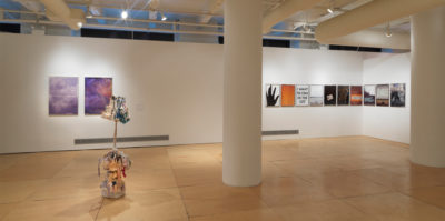 An Installation photograph of the gallery space. On the left side of the wall, there is a diptych of two ink-jet printed images that are about 2 x 3 foot large. Both ink-jet images have a water-color style gradient of purple, white and pink. The gradient is behind three paragraphs of black, bolded text. On the right side of the wall, nine images of the same size and frame are positioned an inch apart from each other. The size of the images are 2x1 foot large. A sculpture is positioned in the center of the space on the floor. Consisting of multiple shapes and layers, a thin piece the size of a stick, is positioned among the middle of the thick layers.