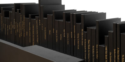 A photograph of a sculpture made of books. The books are stacked vertically in a horizontal line. Every book has black book covers, bindings, and papers. Written vertically on each book binding is a book title in gold paint. Each book size is different in length and height.