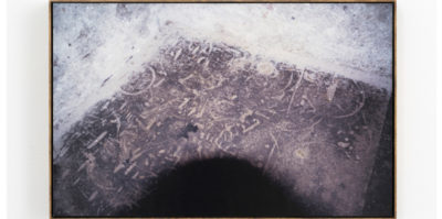 A photograph of an archival pigment print. The print is framed by a small, wooden frame. The print consists of three different colors, layered in a triangular shape. The top layer is white, with black smudge-like accents. The middle layer is grey with white, smudge-like accents. The bottom layer is a dark, black color.