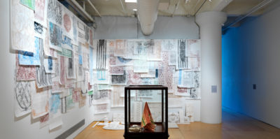 An installation photograph of two gallery walls that meet in a corner. Both walls are covered from ceiling to floor of various drawings on white paper. Each drawing layers over one another. The drawings consist of different shapes and lines in colors oc blue, orange, black, red, and brown. On the floor of the white wall, a horizontal row of unpainted, small, white sculptures are placed. In the center of the space there is a glass vitrine, holding a orange, yellow, and green textile.