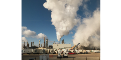 A landscape photograph is taken outside a Citco factory. The landscape of the factory, machinery, cars and containers extends across the lower portion of the photograph. In the middle of the photograph, a big white smoke cloud is coming out of one of the pillars in the factory. It is blended with the clouds on the upper left part of the image. Taking up the entire right side of the landscape is dark gray smoke, covering up the blue sky behind it. There is a red car at the right bottom part of the image.