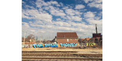A digital photograph that documents a landscape structure beside three sets of train tracks. The wall of the structure is covered in an assortment of graffiti. In the center of the wall, the largest piece of the graffiti is blue text that reads, “The Drug Money”. Peeking behind the graffiti wall is multiple, identical, one-story, brown homes, accompanied with electric poles that stretch into the far distance of the photograph.