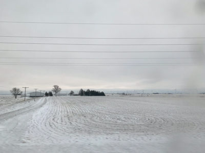 A photograph of a snowy, flat, farming field landscape during the winter season. The entire field is covered in snow, in which the break between sky and land meets in the middle. Houses, trees, and wind turbines can be seen far from the camera’s view, in shades of white and grey.