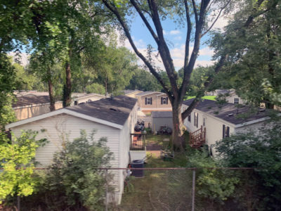 A digital photograph taken behind a metal fence of a backyard of two mobile homes.. The setting is photographed from an elevated point of perspective during a sunny midafternoon. Two sides of the houses are facing each other with a small yard in between them. The shared yard is in the center of the photograph. Three full grown trees with green leaves are planted on the left, center and right side of the houses. Emerging between the tree branches is a clear blue sky with few clouds. The center tree is the largest, extending from the center of the photograph to the top right, center, and left sides, creating a vignette with its leaves and shadow. Beside the trunk of the tree, and the center space of the mobile homes, sits an assortment of bicycles, wooden stairs and a plastic bag.