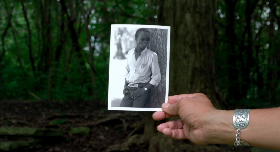 A photograph of a hand holding a black and white polaroid photograph. In the polaroid, there is a middle aged person standing beside a tree. In the background of the image, there is green trees.