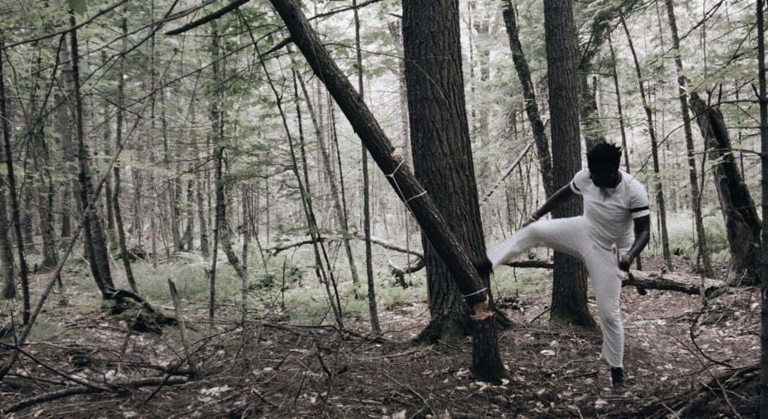 A photograph of a middle aged person in a woods setting. They are wearing all white, and are kicking a tree down.