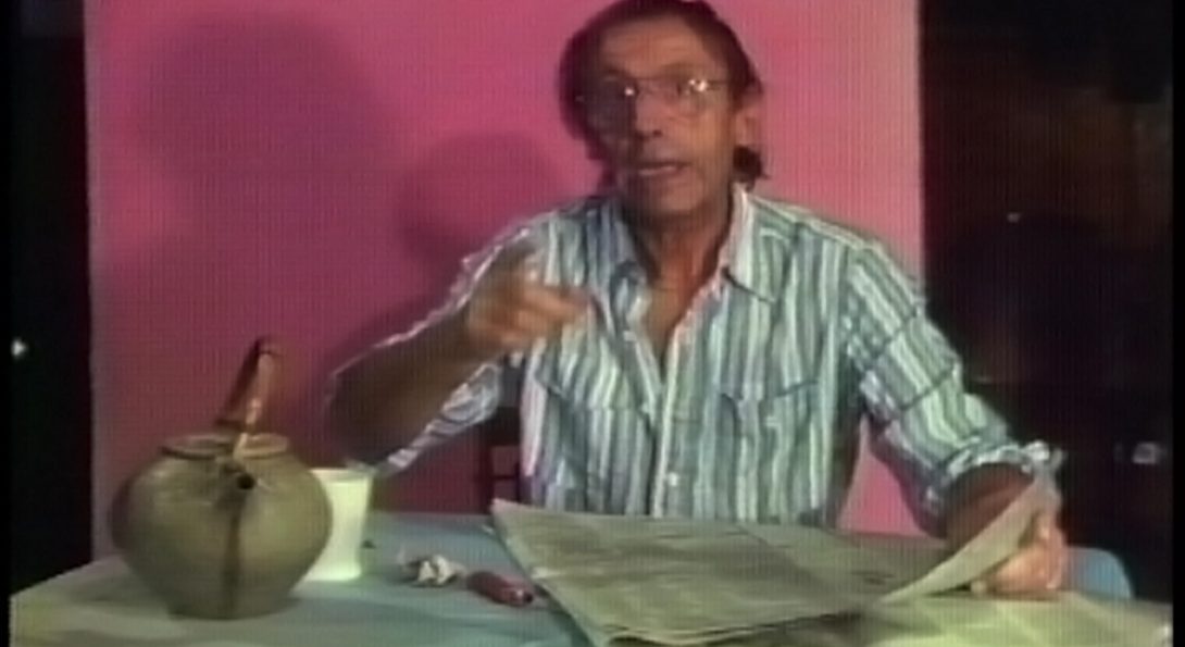 A photograph of a screen still. The screen is paused on an older-aged person, holding the newspaper and talking. They are seated in front of a bright pink background.