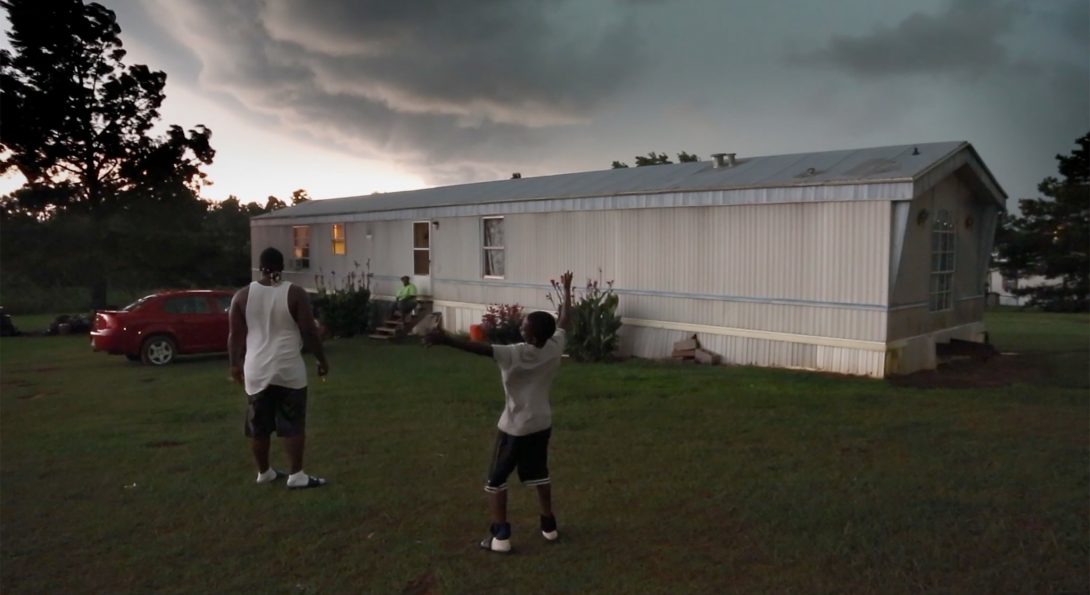 A photograph of a middle-aged person and a younger-aged person are standing in a yard of a mobile home, at dusk.