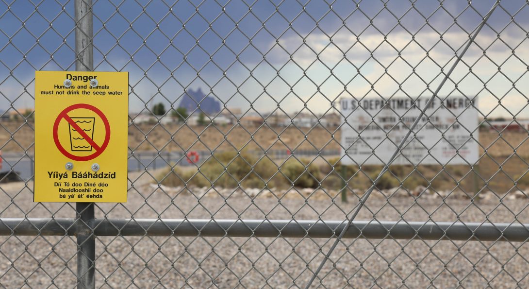 A photograph of a metal fence stands in front of a flat field. A small area of water is situated on the left side. Behind the fence, a large white billboard reads “U.S Department of Energy”. Posted on the fence, a yellow sign with a red circle cross out symbols warns “Danger, humans and animals must not drink water.”