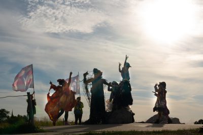 About seven people are silhouetted against a bright day. They are wearing colorful costumes and in the midst of movement.