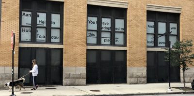 Light brick building with windows lined with white paper with black text. Text reads 