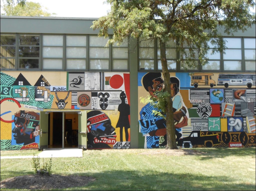 Wide view of a mural on the side of a building. On the right is the mural described above and on the left is a continuation of that mural with more grid-style images along with a silhouetted figure and two large black faces on either side of the building’s door, facing away from the two sides of an open double door.