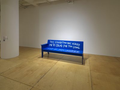 Blue bench with white text that reads: This exhibition has asked me to stand for too long. Sit if you agree.