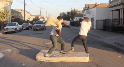 One Black man and one white man are engaged in a pillow fight in the center of an urban street. They are balanced on a tattered mattress as a vehicle drives toward them.