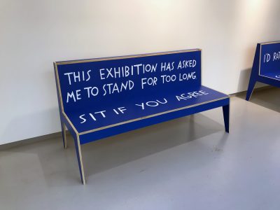 Blue wooden bench in a gallery with text painted on it. The back of the bench reads, 