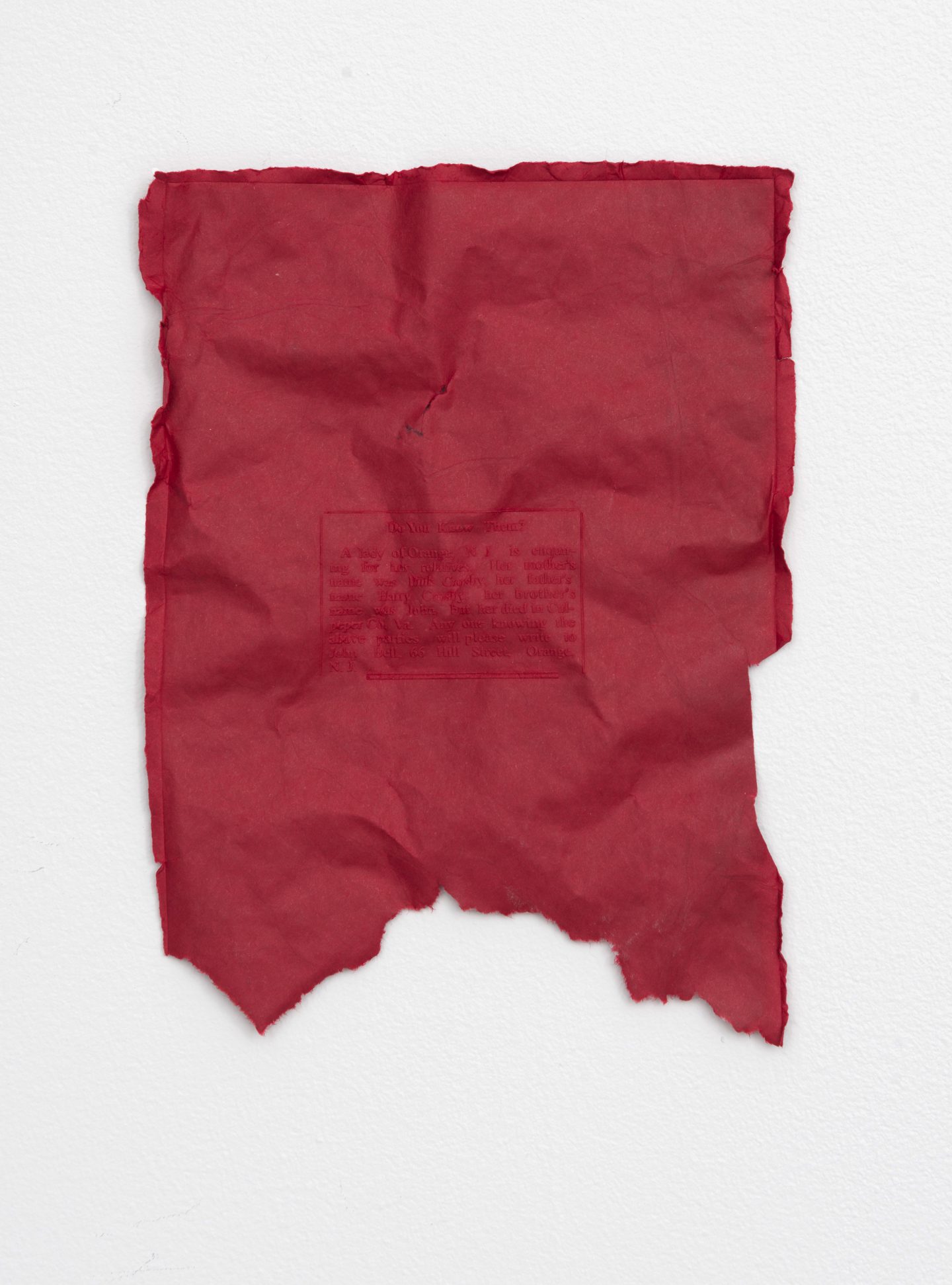 On a white wall, a single piece of crumpled and torn red paper.