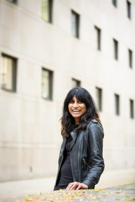 A young woman in a leather jacket smiles at the camera in front of a concrete building.