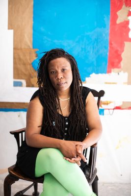 A black person with long dreadlocks sits on a chair and looks into the camera.