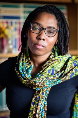 A young black woman wearing a green scarf and glasses looks at the camera with a neutral face.