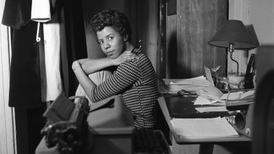 Black and white image of a black woman next to a desk looking at the camera.