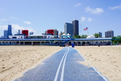 A view of the Chicago skyline from the middle of North Avenue Beach. In the center of the beach is a soft blue plastic ramp, with sand on either side. At the end of the ramp is a building that looks like a boat, with white walls, porthole windows, and bright red steam pipes. An American flag blows in the wind against a pale blue sky.