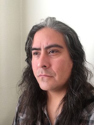 A medium-skinned person with long dark hair looks just to the side of the camera.
