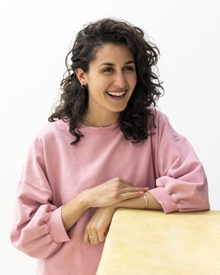 A medium skinned person with brown curly hair and wearing a pink top. They lean on a table and smile, looking to the right of the viewer.