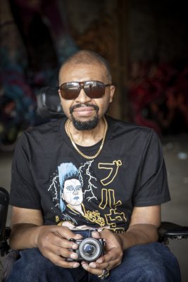 A black man in a black shirt and sunglasses looks directly at the viewer.