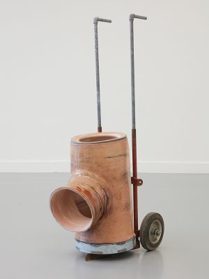 A sculpture in a white walled space. The object is made of a tube with a circular opening in the front and two pipes stretched up the back. The object is on wheels.