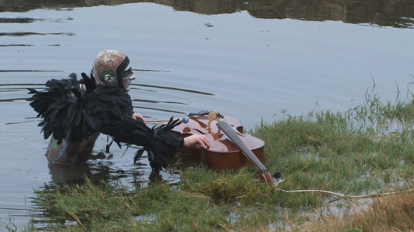 A figure wearing feathers and a helmet stands partly submerged in water and plays a cello that floats in the same water.
