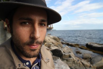 Against a blurry background of rocks and ocean, a close-up of the artist Carmen Papalia, an olive-skinned man with brown eyes, a dark, close-trimmed beard, and a gray hat.