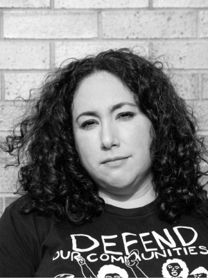 Black and white photo of a person with light skin and curly dark hair. Their shirt is black and says in white letters, 