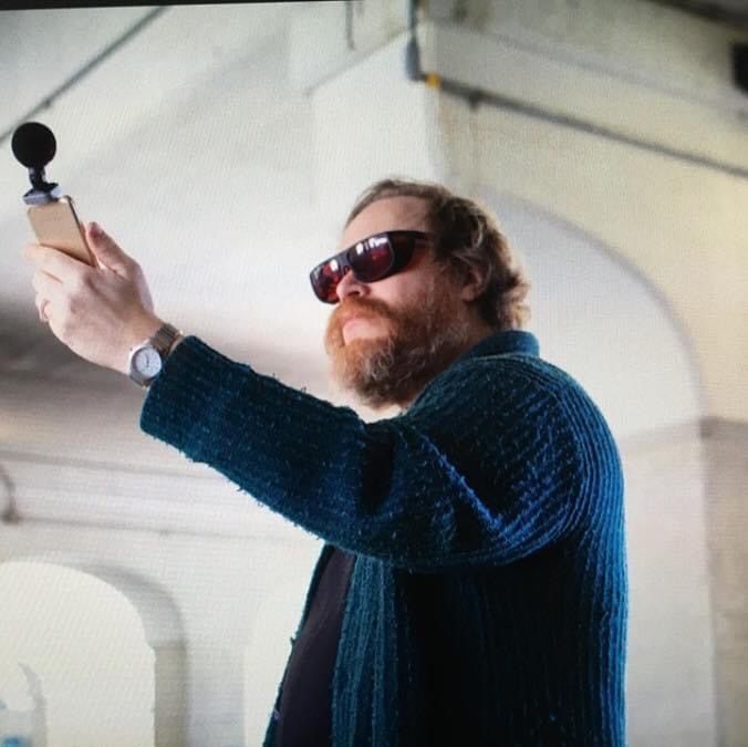 A white man with sunglasses on wearing a blue sweater holds up a phone with a microphone attached.