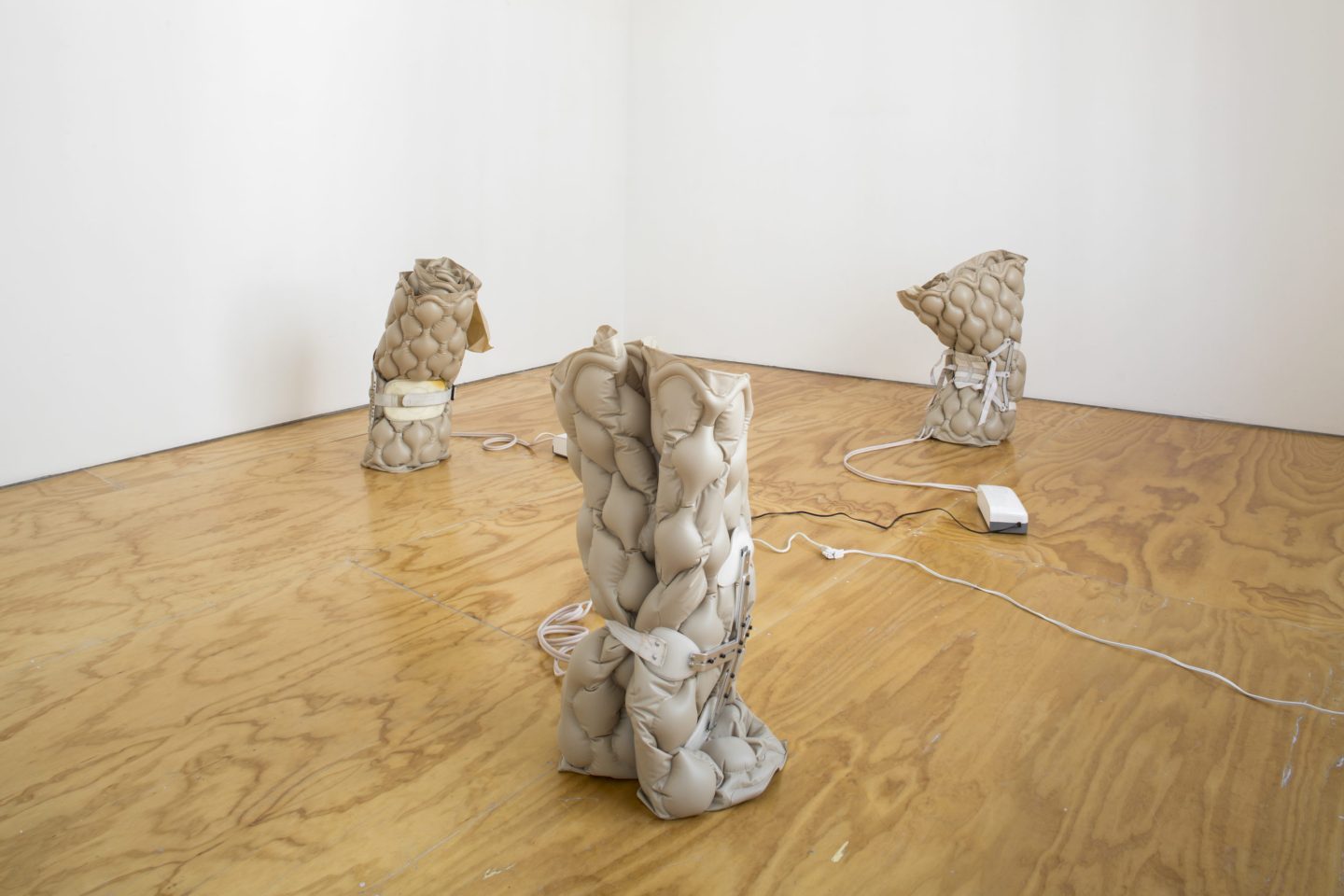 Three sculptures sit on the wood floor. They are made of lumpy white pressure pads encased in braces and are connected by cords.